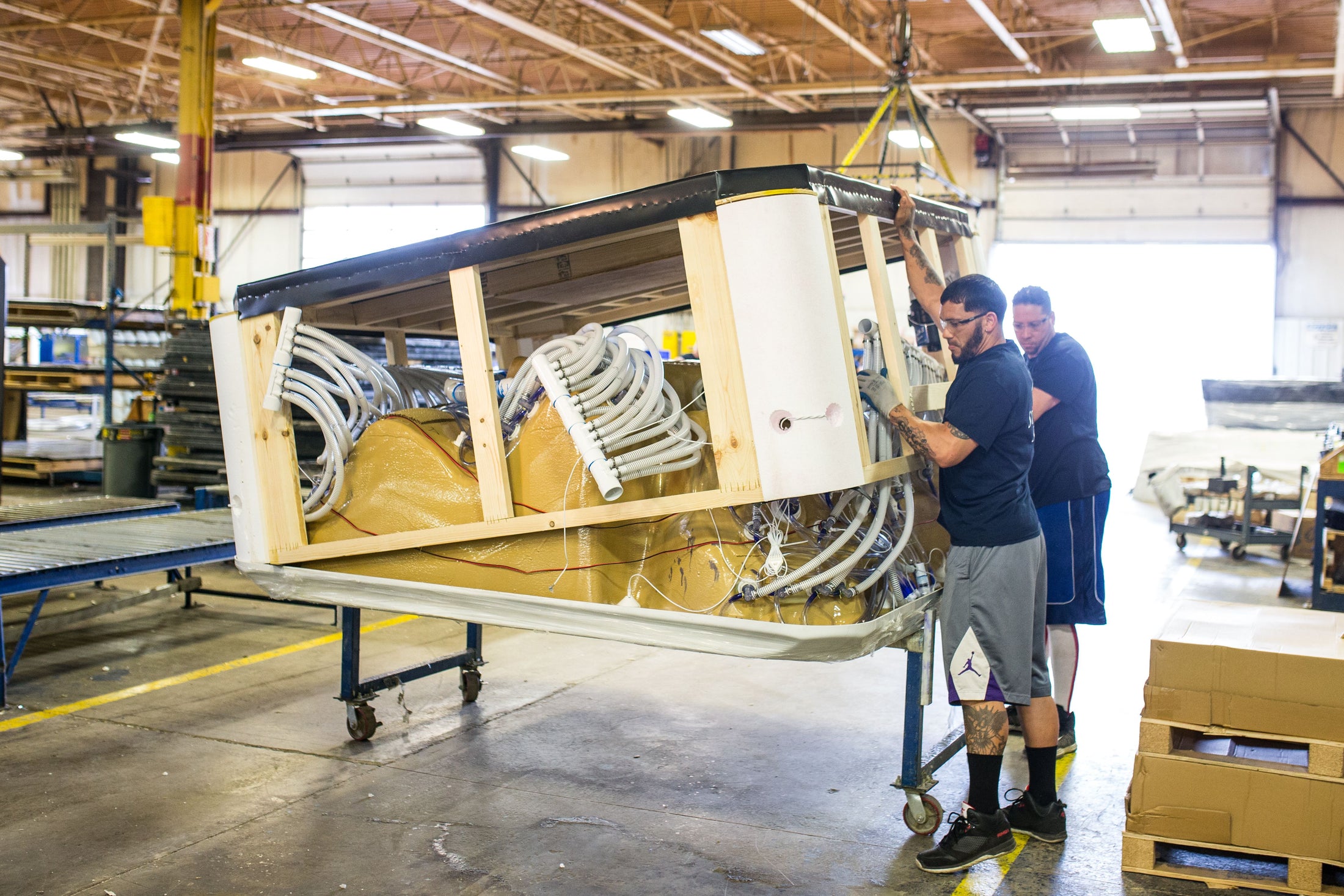A hot tub being built in a factory with two workers standing beside it and handling parts or tools. The scene shows a real industrial environment and people, so this is Lifestyle rather than Product/Packaging.